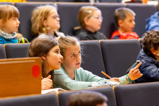 Kinder die in einem Hörsaal sitzen - zwei Mädchen sind im Vordergrund und schauen in die Ferne