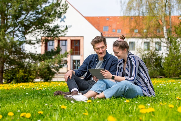 Zwei Studierende sitzen auf einer Wiese mit einem Tablet, im Hintergrund ist ein Haus zu sehen