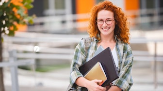 Eine lächelnde rothaarige Doktorandin vor einer Hochschule