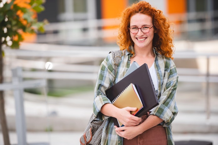 Eine lächelnde rothaarige Doktorandin vor einer Hochschule