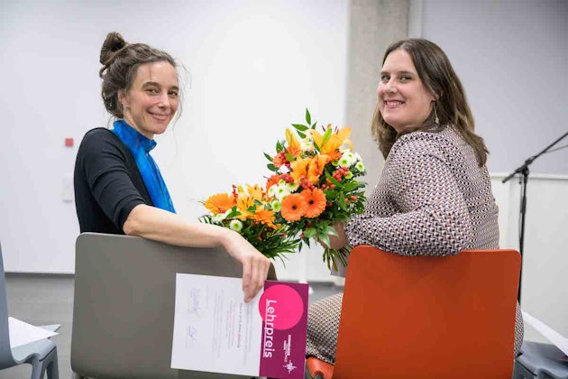 Eröffnung der EH Freiburg - Zwei Frauen sitzen auf einem Stuhl und drehen sich Richtung Kamera, beide halten einen Blumenstrauß in der Hand
