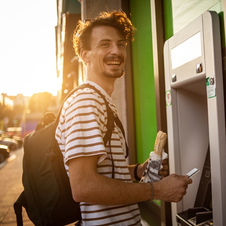 Ein junger Mann am Geldautomat freut sich über sein Gehalt im öffentlichen Dienst