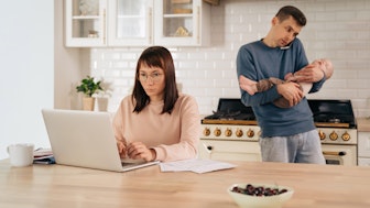 Job und Familie: Eine Frau am Laptop und ein Mann mit Handy am Ohr und Kind auf dem Arm in der Küche