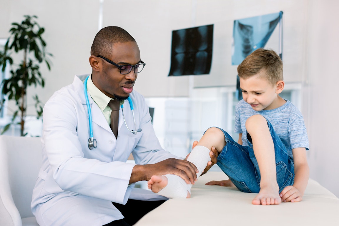 Ein dunkelhäutiger Arzt untersucht den eingegipsten Fuß eines Jungen