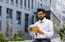 Ein junger Mann vor einem Unigebäude zieht lächelnd ein Schreiben aus einem Briefumschlag