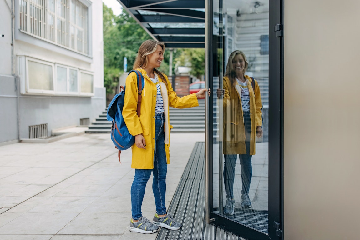 Eine Studentin in gelbem Regenmantel öffnet eine Tür zur Uni, in der sie sich spiegelt