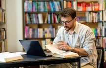 Ein junger Mann am Laptop in einer Bibliothek will aus einem Buch zitieren