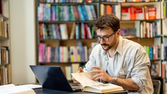 Ein junger Mann am Laptop in einer Bibliothek will aus einem Buch zitieren